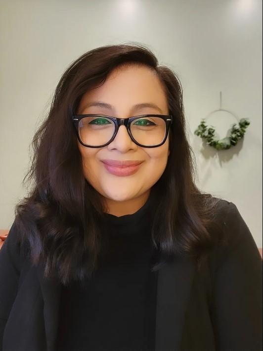 Therapist Leslie Martinez smiling at the camera wearing glasses and a black shirt with a black blazer and a plant in the background